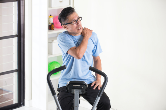 Asian Man Experiencing Shoulder Pain On Exercise Bike